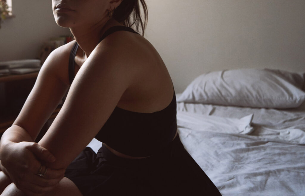 Valeria Cisneros a model with a contemplative expression sits on a bed in a dimly lit room wearing a black tank top and skirt with a bedside table and a window in the background.
